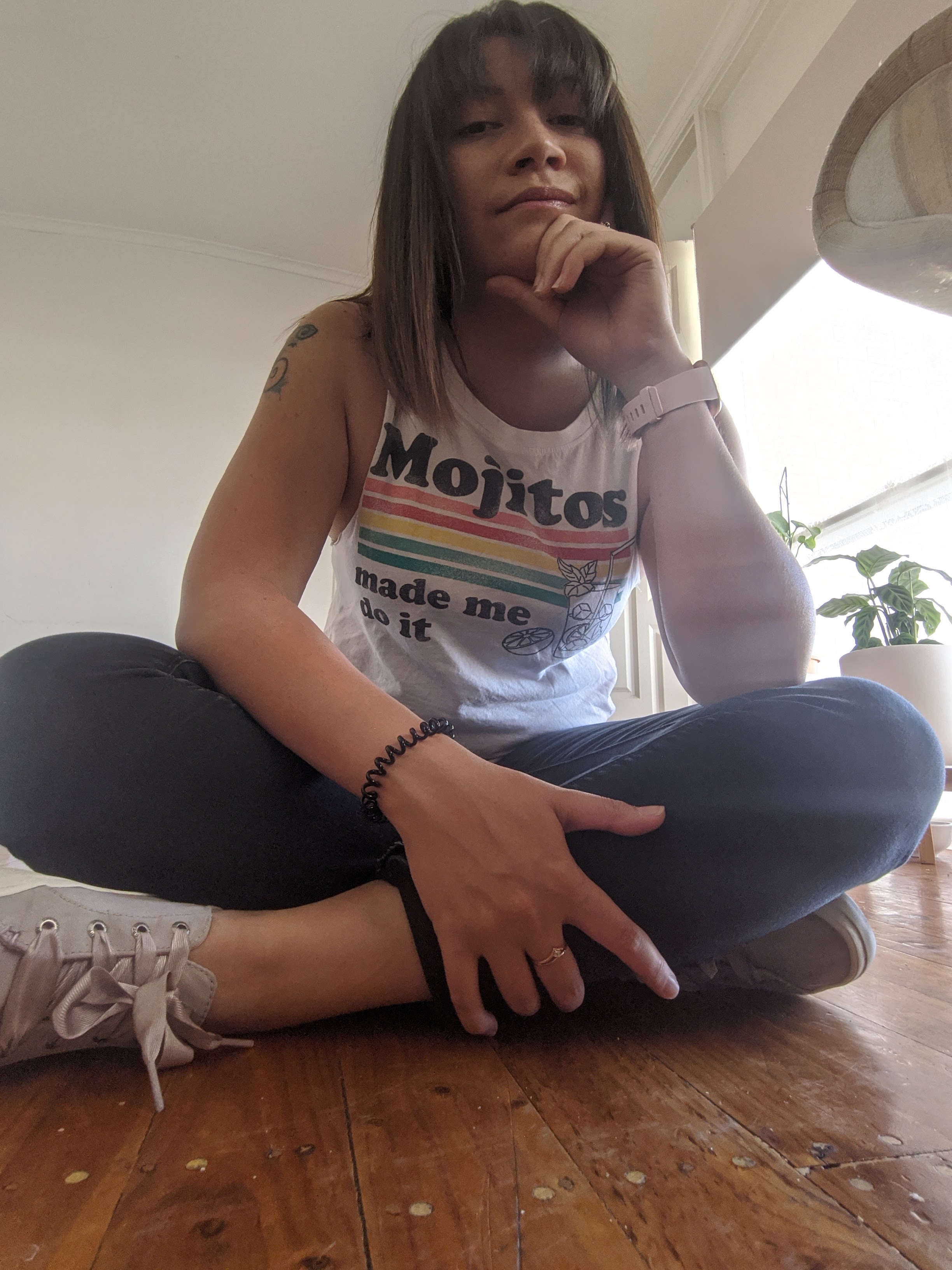 A light-skinned woman with long brown hair sits cross-legged on the floor with one hand under her chin. She is wearing white sneakers, black sweatpants, and a white tank top that reads 'Mojitos made me do it'. She is half-smiling at the camera, which is looking up at her from the floor.